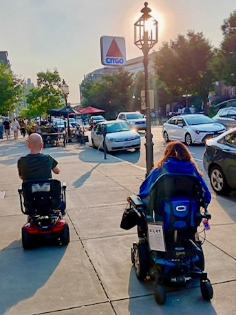 Two people using a mobility scooter and a power wheelchair travel side by side on a Boston sidewalk, navigating city life together.