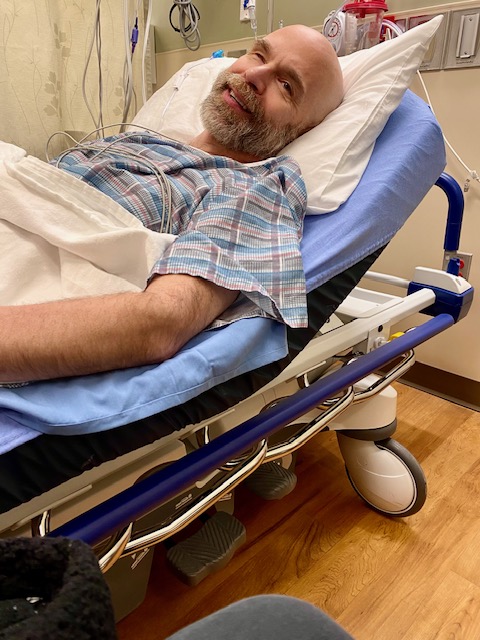 Dan resting on a hospital gurney before surgery, smiling with calm reassurance while waiting to be taken in.