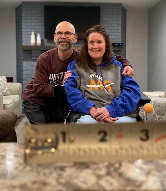 Dan and Jennifer Digmann sitting together at home during post-surgery recovery, with a measuring tape visible in the foreground.