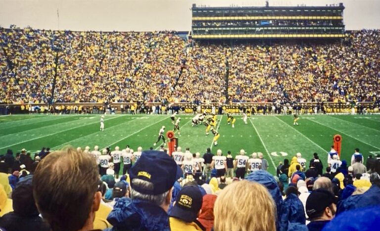 View of Michigan Stadium, known as the Big House, during a University of Michigan football game with the crowd and marching band visible on the field.