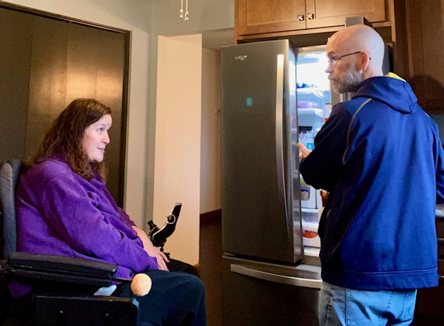 Man with multiple sclerosis stands at an open refrigerator while his partner watches, capturing a quiet everyday moment of forgetfulness and life with MS
