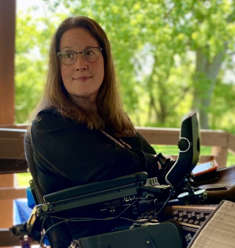 Jennifer Digmann sits on her covered deck in her wheelchair, looking thoughtfully toward the yard surrounded by summer greenery.