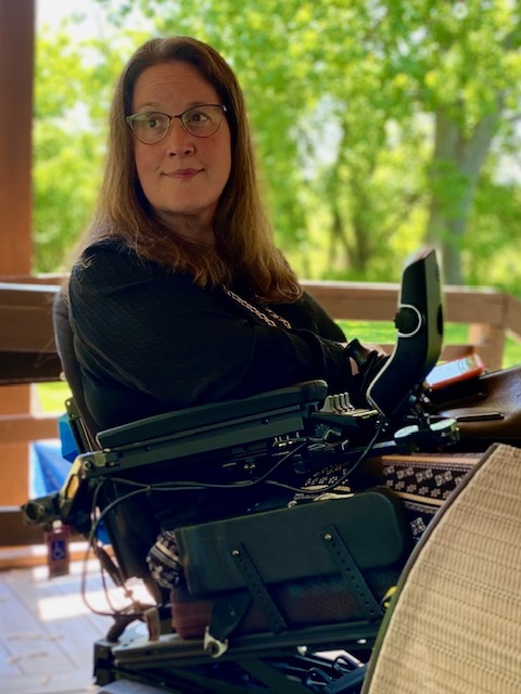 Jennifer Digmann sits on her covered deck in her wheelchair, looking thoughtfully toward the yard surrounded by summer greenery.